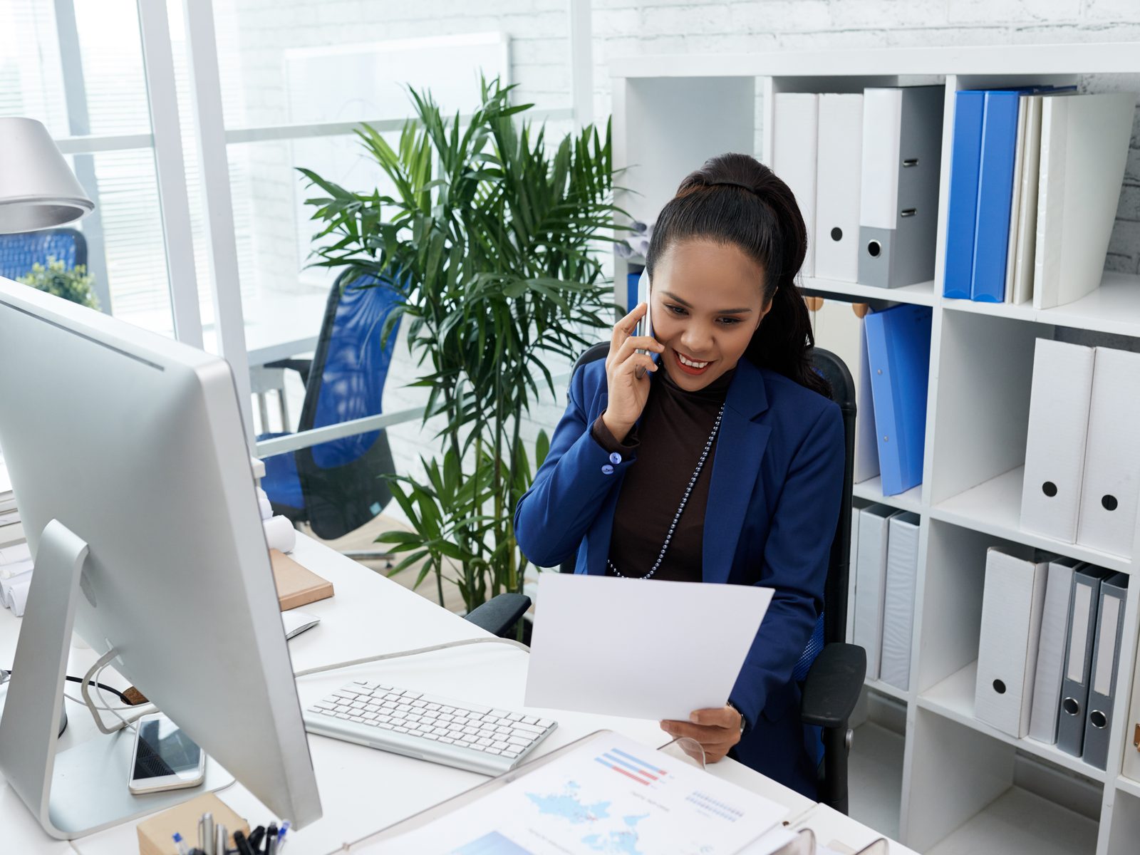 Administrative services manager on the phone at her desk, reviewing a document beside shelves of office binders
