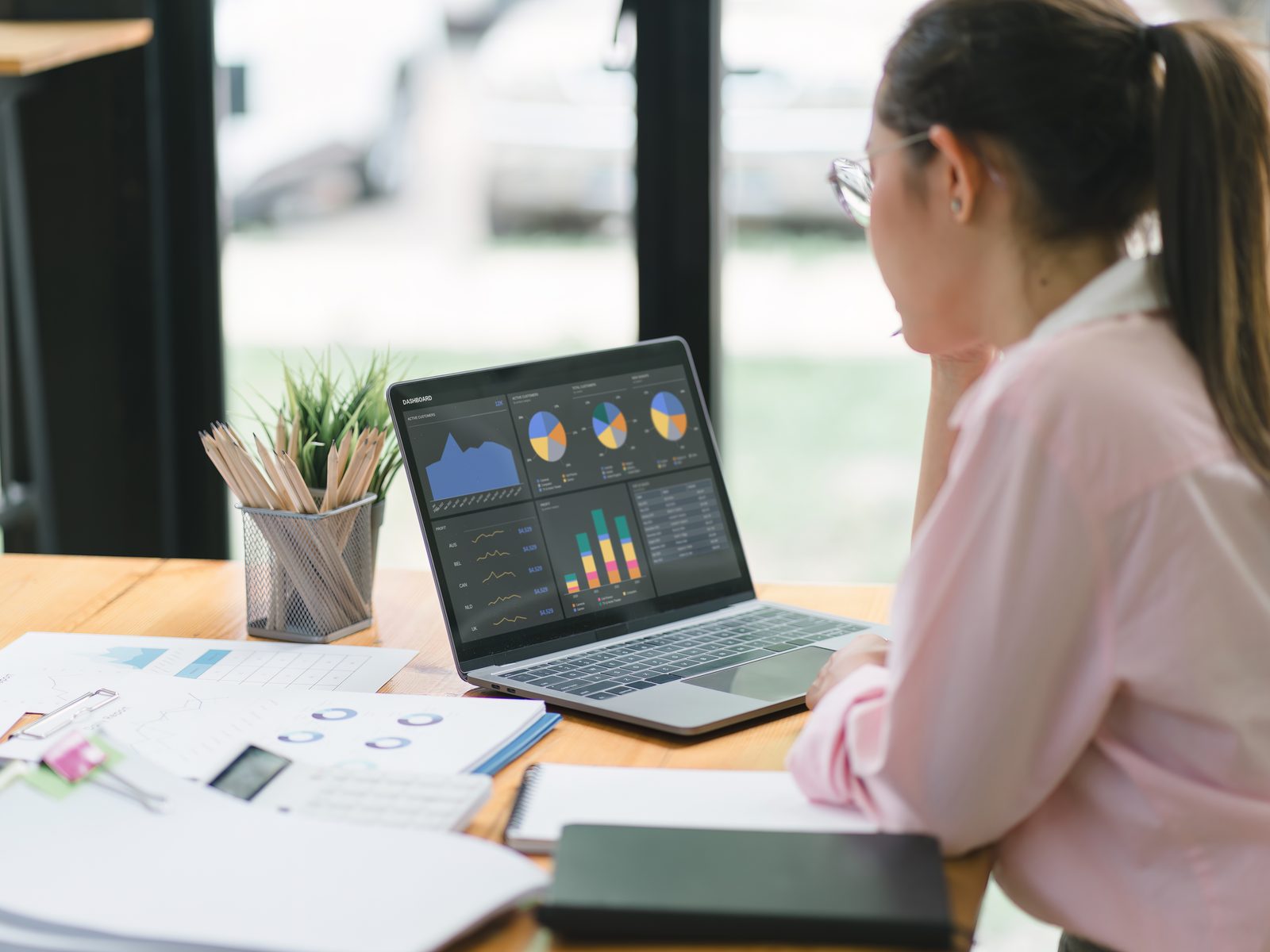 Management analyst studying charts on a laptop dashboard with printed reports and a notebook on the desk