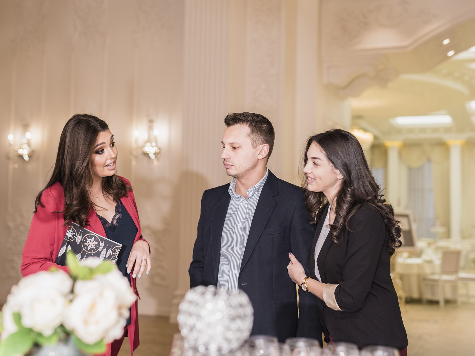 Event planner holding a menu folder and walking a couple through a banquet hall with set tables and flowers