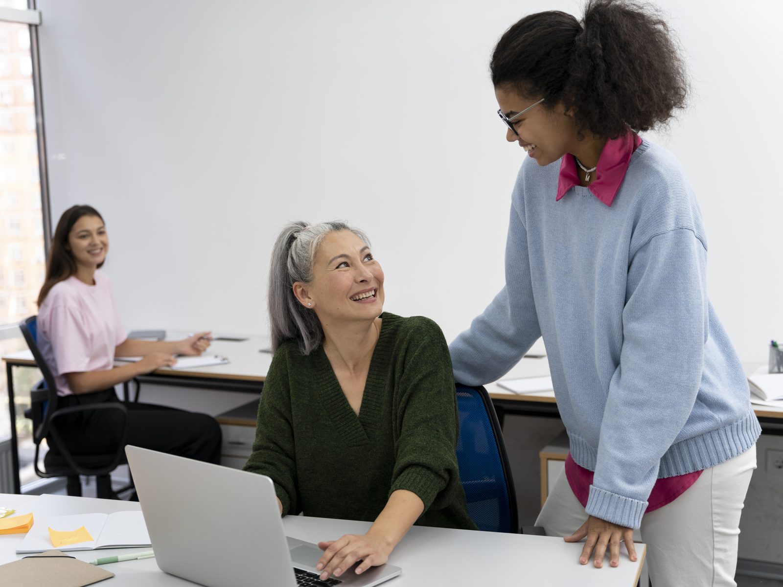 Social and human service assistant leaning over to help a woman working at a laptop in a shared office