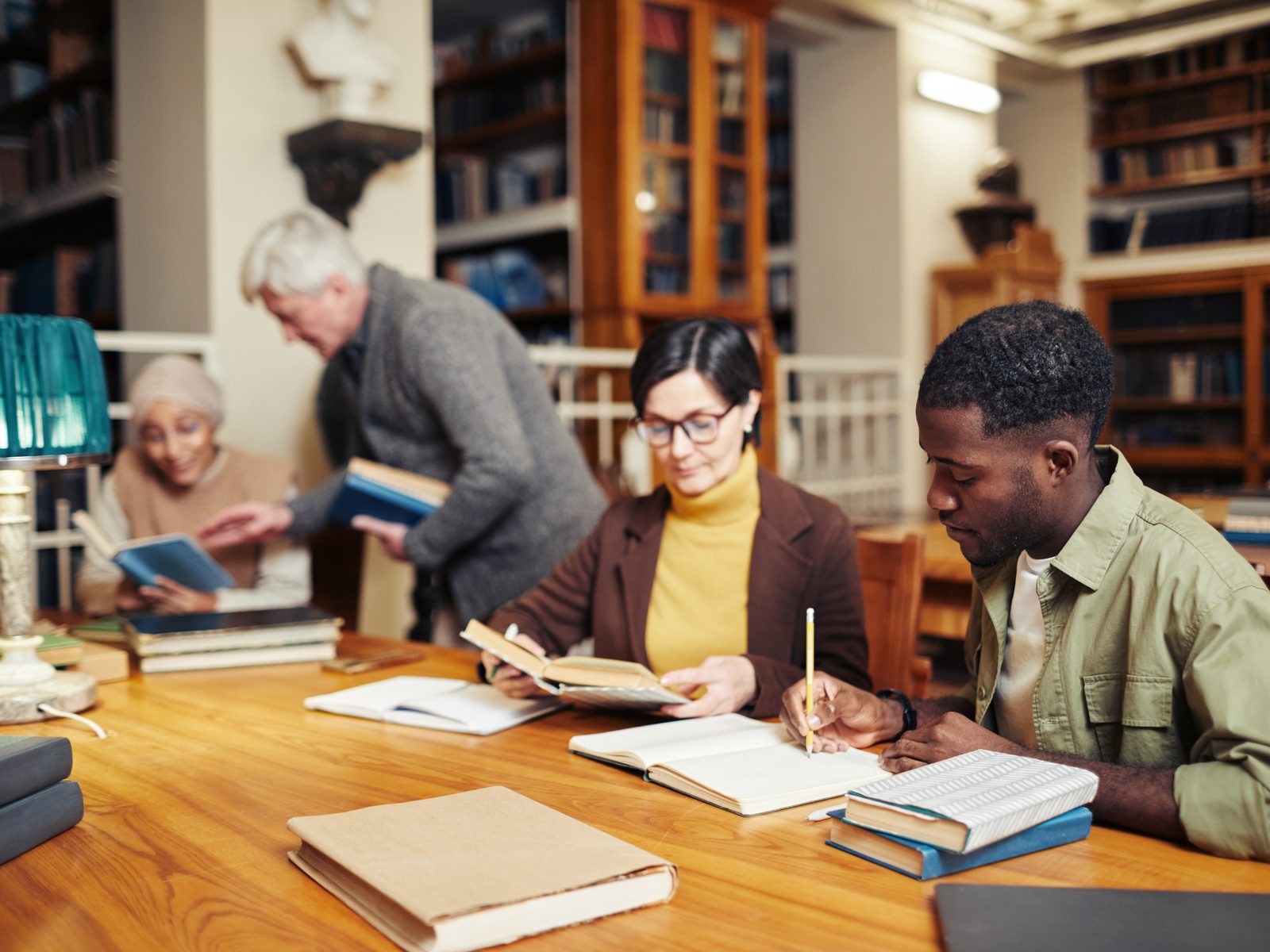 Adults of different ages reading and taking notes in a library reading room, as in a doctoral program in liberal studies and humanities