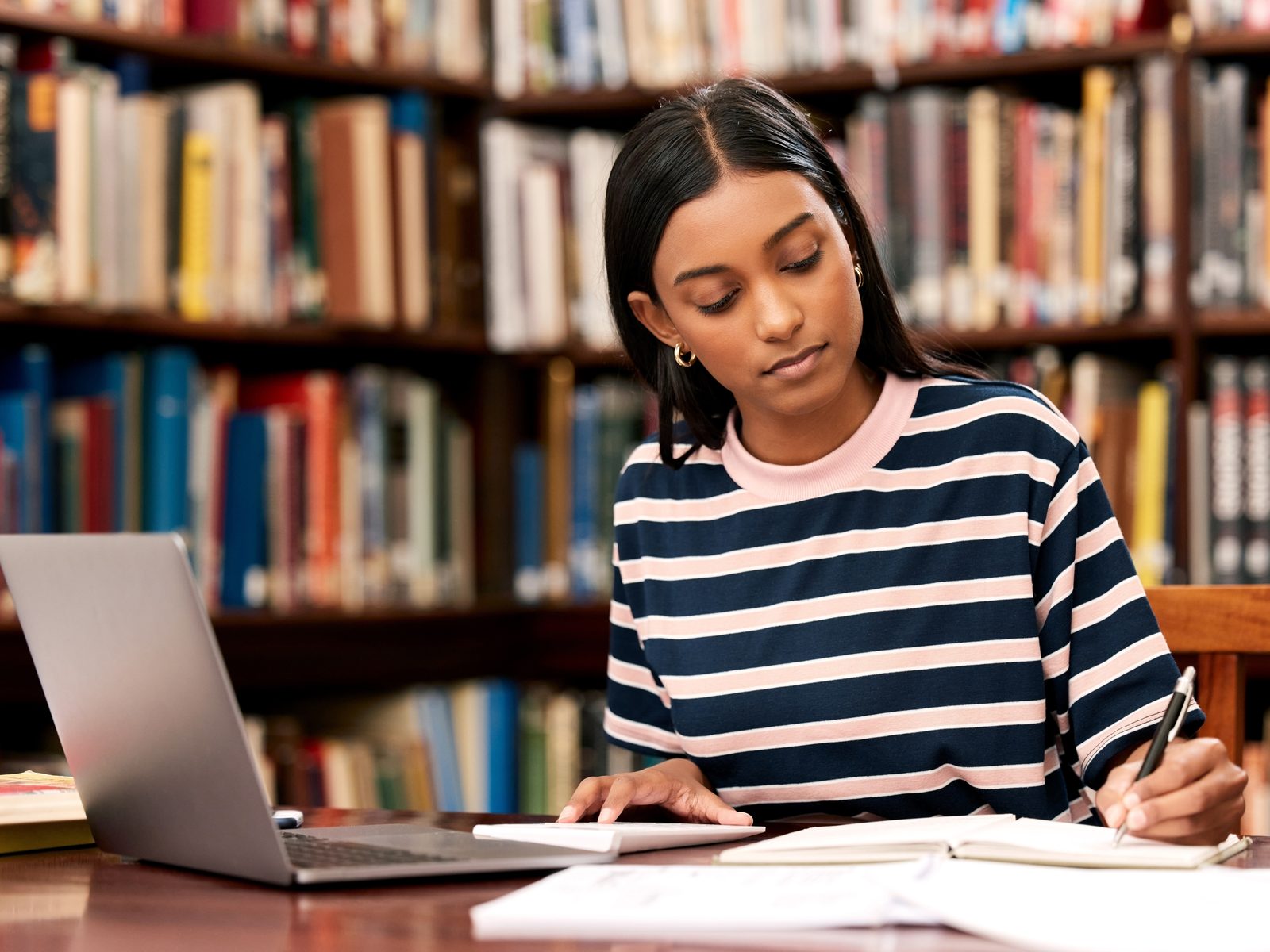 Student taking notes beside a laptop in a library while studying for an online associate degree in general studies